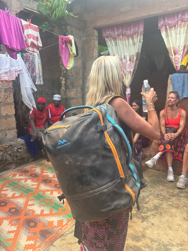 A tourist woman carrying a trekarius backpack with orange and blue straps, standing in a rustic outdoor village courtyard in zanzibar with people and laundry visible in the background.