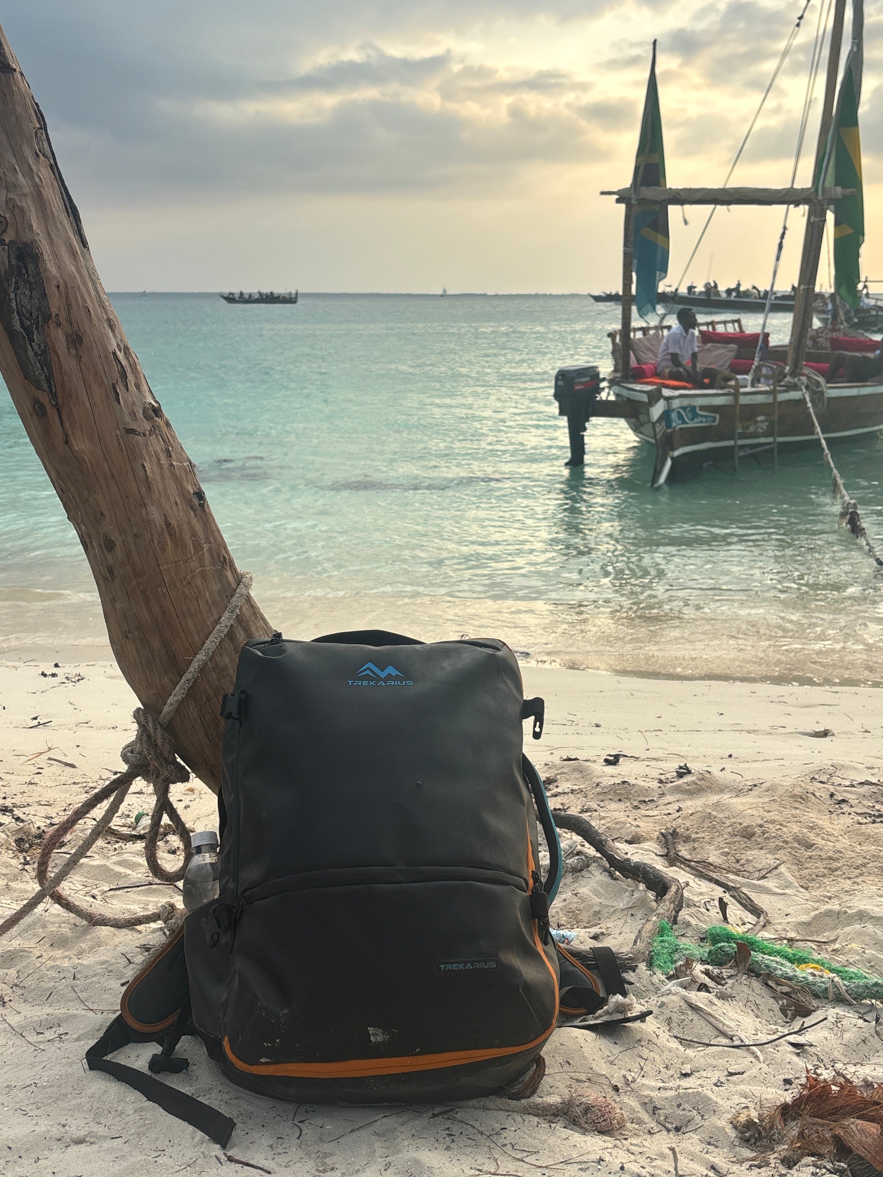 TREKARIUS 35L backpack resting on a sandy beach next to a wooden pole with a tropical lagoon in the background