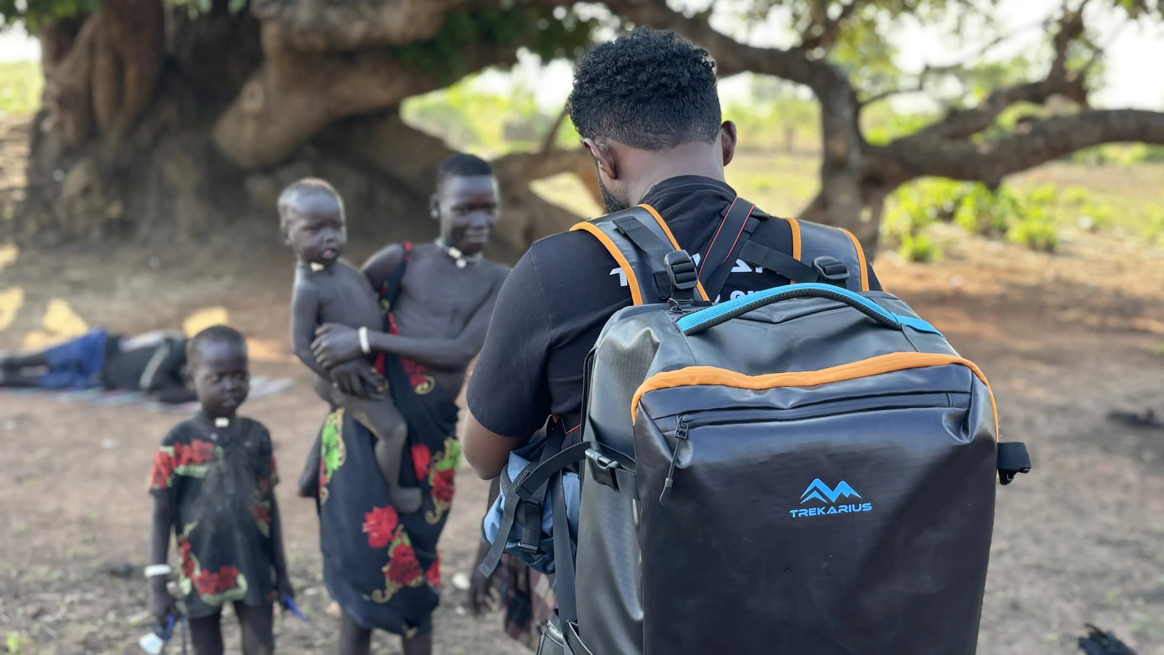A man wearing a trekarius backpack stands outdoors under a large tree, facing three local children in a rural setting.